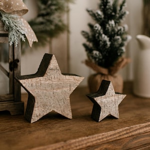 May include: Two rustic wooden star decorations, one large and one small, with a natural wood grain finish and dark edges. The stars are placed on a wooden surface, suggesting a holiday or seasonal theme.