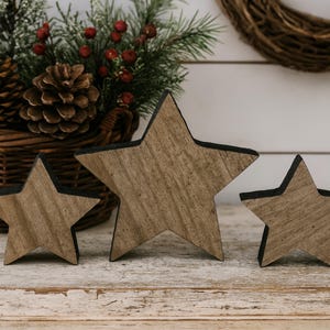 May include: Three wooden star decorations with a rustic, weathered appearance. The stars vary in size and have a natural wood grain finish with black edges. A basket with pinecones and greenery is in the background.