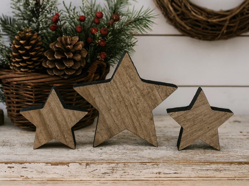 May include: Three wooden star decorations with a rustic, weathered appearance. The stars vary in size and have a natural wood grain finish with black edges. A basket with pinecones and greenery is in the background.