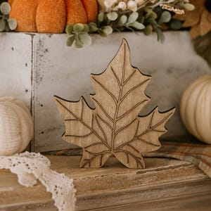 May include: A wooden maple leaf decoration with detailed etched lines, set against a backdrop of fall-themed decor, including pumpkins and sunflowers. The leaf is a light brown color.