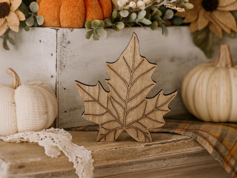 May include: A wooden maple leaf decoration with detailed etched lines, set against a backdrop of fall-themed decor, including pumpkins and sunflowers. The leaf is a light brown color.