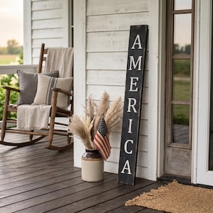 May include: A rustic porch scene features a wooden rocking chair with pillows and a blanket. A black sign with the word "AMERICA" in white lettering leans against the wall. A vase with pampas grass and an American flag completes the patriotic decor.