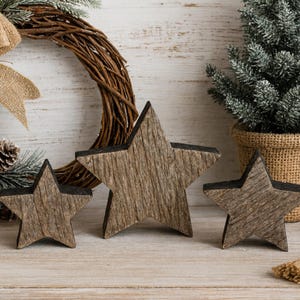 May include: Three wooden star decorations with a rustic, weathered appearance. The stars are various sizes and have a dark edge. A wreath, a small Christmas tree in a burlap pot, and other holiday decorations are in the background.