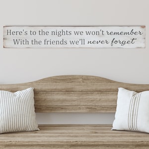 May include: A wooden bench with two striped pillows and a decorative sign. The sign reads "Here's to the nights we won't remember With the friends we'll never forget."