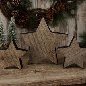 May include: Three wooden star decorations with a rustic, weathered appearance. The stars vary in size and have a natural wood grain finish with black edges. They are arranged on a wooden surface, suggesting a holiday or seasonal theme.