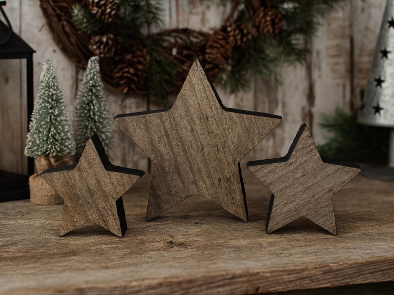 May include: Three wooden star decorations with a rustic, weathered appearance. The stars vary in size and have a natural wood grain finish with black edges. They are arranged on a wooden surface, suggesting a holiday or seasonal theme.