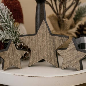 May include: Three wooden star decorations with a rustic, weathered appearance. The stars vary in size and have a dark border. They are displayed on a white, round tray, with festive decor in the background, including a gnome and pinecone.