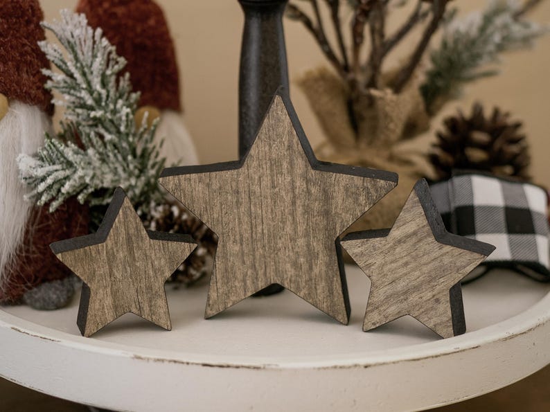 May include: Three wooden star decorations with a rustic, weathered appearance. The stars vary in size and have a dark border. They are displayed on a white, round tray, with festive decor in the background, including a gnome and pinecone.