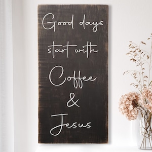May include: A rectangular, dark brown wooden sign with white cursive text that reads "Good days start with Coffee & Jesus." The sign is displayed near a vase of dried flowers.