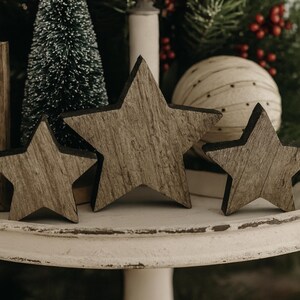 May include: Three wooden star decorations with a rustic, weathered appearance. The stars are arranged on a white, tiered stand, with a small Christmas tree and decorative ball in the background. The stars vary in size.