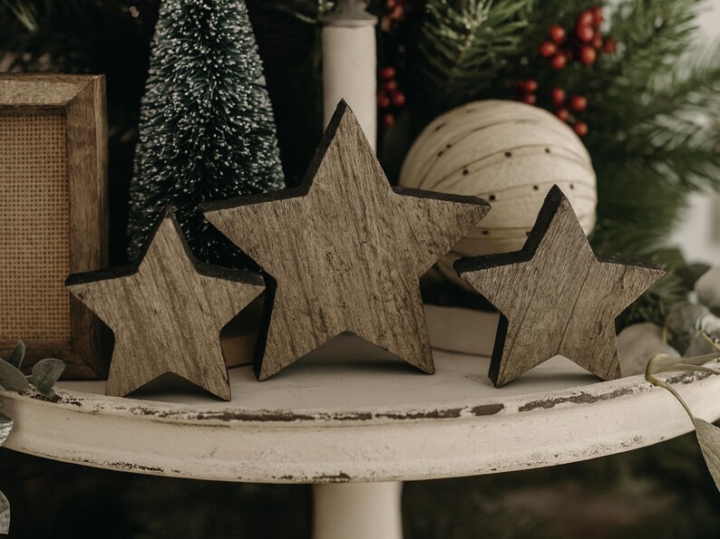 May include: Three wooden star decorations with a rustic, weathered appearance. The stars are arranged on a white, tiered stand, with a small Christmas tree and decorative ball in the background. The stars vary in size.