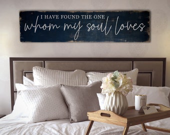 Puede incluir: Un letrero de madera azul marino con el texto "I HAVE FOUND THE ONE whom my soul loves" en escritura blanca cuelga sobre una cama con almohadas blancas y una bandeja de madera con un jarrón de flores. El letrero tiene un aspecto envejecido.
