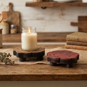 May include: A lit candle in a glass jar sits on a wooden coaster with a floral design. A second coaster with a rose design is next to it. The coasters are on a wooden table, with books and kitchen items in the background.