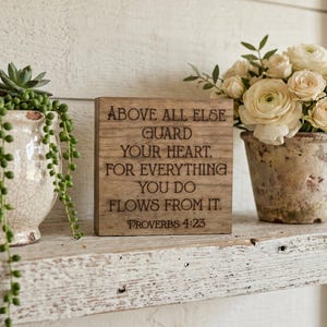 May include: A wooden sign with the text "ABOVE ALL ELSE GUARD YOUR HEART. FOR EVERYTHING YOU DO FLOWS FROM IT. PROVERBS 4:23" sits on a distressed white shelf. A potted plant with trailing green vines and a vase of white flowers are also on the shelf.