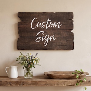 May include: A rustic wooden sign with the words "Custom Sign" in white script. The sign is mounted on a beige wall above a wooden shelf. The shelf holds a white pitcher, a glass jar of flowers, and a wooden bowl.