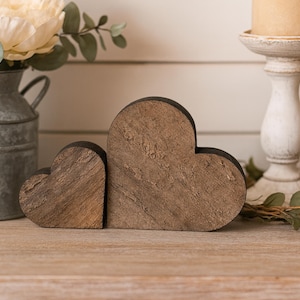 May include: Two wooden heart-shaped decorations with a rustic finish. The hearts are different sizes and stand on a wooden surface. A metal vase with flowers and a white candle holder are in the background.