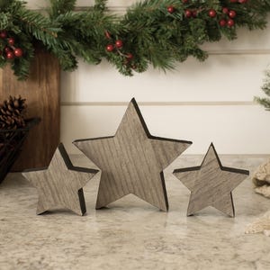 May include: Three wooden star decorations in varying sizes. The stars have a gray wood grain appearance with dark edges. The stars are arranged on a speckled countertop, with a Christmas garland and small tree in the background.