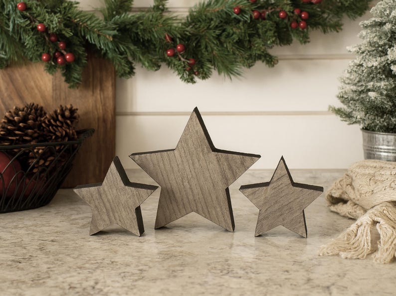 May include: Three wooden star decorations in varying sizes. The stars have a gray wood grain appearance with dark edges. The stars are arranged on a speckled countertop, with a Christmas garland and small tree in the background.