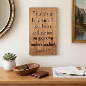 May include: A wooden sign with the text "Trust in the Lord with all your heart, and lean not on your own understanding. Proverbs 3:5." Also, a succulent in a white pot, a wooden bowl with keys, a brown wallet, and a magazine.
