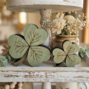 May include: Two wooden shamrock decorations, one large and one small, with green leaves and black outlines, sit on a distressed white tray. The shamrocks have detailed vein patterns. A small vase of flowers is in the background.