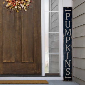 May include: A dark wooden door with a wreath and a blue sign that says "PUMPKINS" in white lettering. The sign has an arrow pointing to the left. The door has a brass handle and lock.