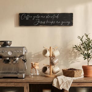May include: A coffee station setup with a stainless steel espresso machine, a wooden mug tree holding six beige mugs, and a small potted olive tree. A sign above reads "Coffee gets me started, Jesus keeps me going."