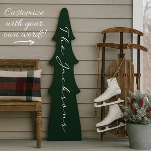 May include: A dark green wooden Christmas tree-shaped sign with the name "The Jacksons" in white script. A wooden bench with a plaid pillow, a vintage wooden sled, and white ice skates complete the holiday scene. Text reads "Customize with your own words!"