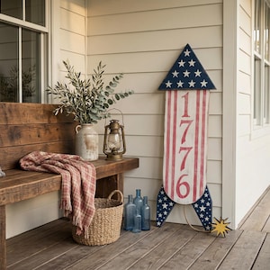 May include: A rustic outdoor scene featuring a wooden bench with a patterned pillow and a red plaid blanket. A decorative rocket with red and white stripes and the number 1776 stands nearby. Additional decor includes a woven basket, blue bottles, and a vintage lantern.