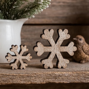 May include: Two wooden snowflake ornaments, one large and one small, sit on a rustic wooden surface. The snowflakes have a natural wood grain finish. A white pitcher with greenery and a small decorative bird are in the background.