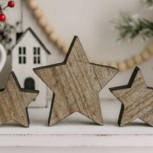 May include: Three wooden star decorations with a rustic, weathered appearance. The stars vary in size and have a natural wood grain finish with dark edges. They are arranged on a white surface, suggesting a holiday or seasonal theme.