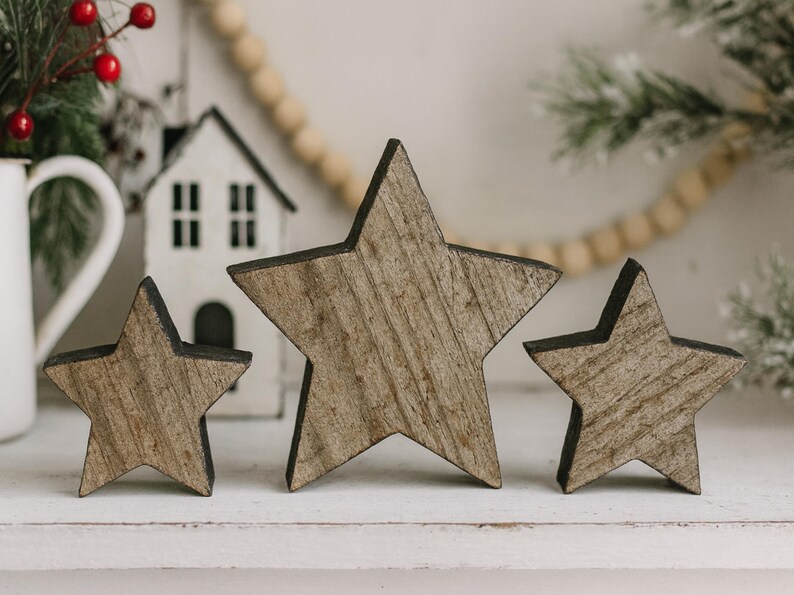 May include: Three wooden star decorations with a rustic, weathered appearance. The stars vary in size and have a natural wood grain finish with dark edges. They are arranged on a white surface, suggesting a holiday or seasonal theme.