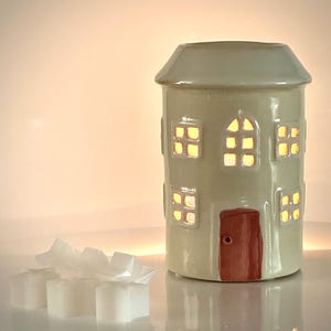 May include: A ceramic house-shaped oil warmer in a pale green colour, with multiple illuminated windows and a red door. The warmer has a chimney and is accompanied by several white wax melts.