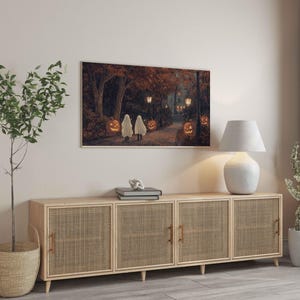 May include: A light wood cabinet with three woven rattan doors. The cabinet is sitting on a light wood floor. A white lamp with a white shade is on the left side of the cabinet. A potted plant is on the right side of the cabinet. A framed picture of a Halloween scene is hanging on the wall above the cabinet.