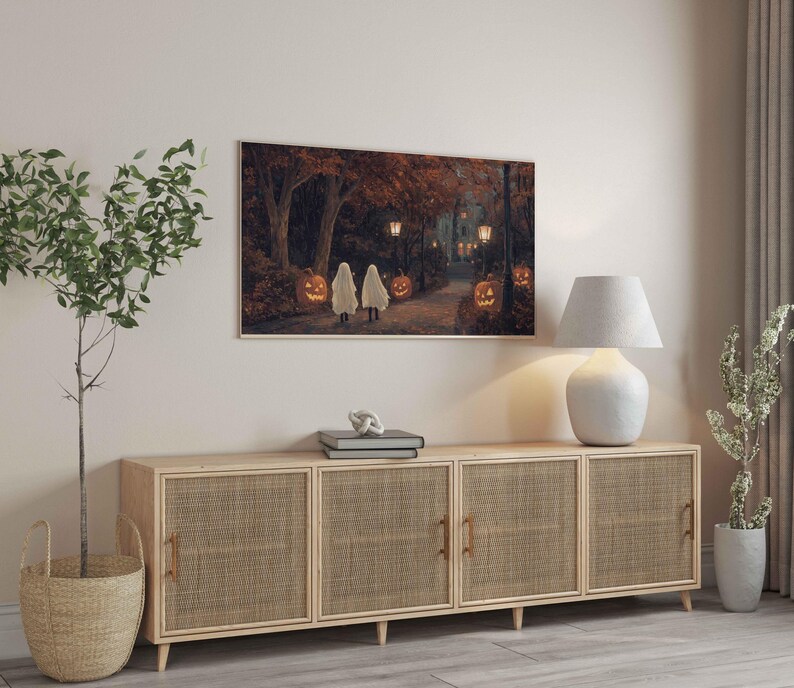 May include: A light wood cabinet with three woven rattan doors. The cabinet is sitting on a light wood floor. A white lamp with a white shade is on the left side of the cabinet. A potted plant is on the right side of the cabinet. A framed picture of a Halloween scene is hanging on the wall above the cabinet.