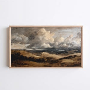 May include: A framed landscape painting depicts rolling hills under a dramatic sky filled with fluffy, light-colored clouds. The artwork uses a muted color palette of browns, tans, and grays, creating a vintage aesthetic.