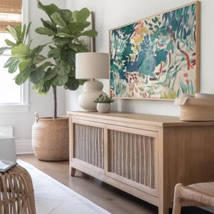 May include: A light wood cabinet with woven panels, topped with a lamp, small plant, and a framed abstract painting. A large potted plant and a woven basket are also in the room, creating a natural, neutral aesthetic.