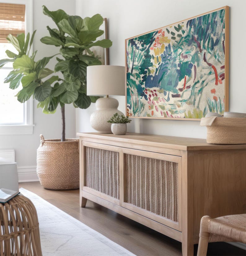 May include: A light wood cabinet with woven panels, topped with a lamp, small plant, and a framed abstract painting. A large potted plant and a woven basket are also in the room, creating a natural, neutral aesthetic.