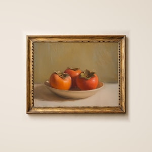 Könnte beinhalten: Ein Ölgemälde in einem goldfarbenen, verzierten Rahmen zeigt drei leuchtend orangefarbene Kakis in einer flachen, cremeweißen Schale. Der Hintergrund ist in einem gedämpften, neutralen Olivgrün gehalten, während die Fläche unter der Schale cremefarben ist.
