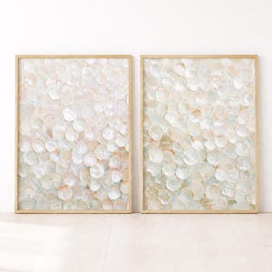 Op de afbeelding: Een paar rechthoekige kunstprints ingelijst in licht hout. Elke print toont een dicht, gelaagd patroon van cirkelvormige, iriserende, schelpachtige vormen in zachte tinten crème, lichtroze, beige en gedempt saliegroen met een parelmoerglans.