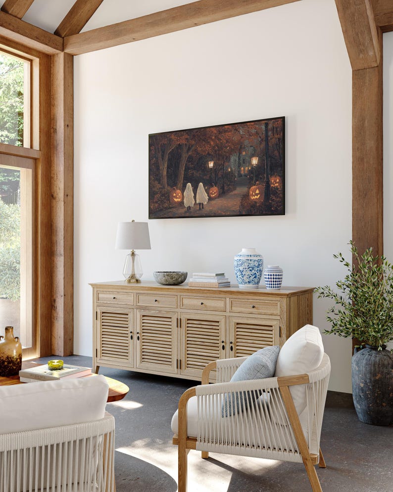 May include: A living room with a light wood cabinet, a white armchair, and a framed painting of children trick-or-treating in a forest. The painting features a dark background with glowing pumpkins and lanterns.