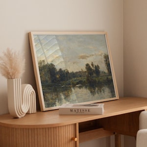 May include: A framed landscape painting of a river scene with trees and a cloudy sky. The painting is hanging on a wall above a wooden desk with a book titled "MATISSE" on top of it.