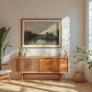 May include: A wooden cabinet with two doors and a drawer, featuring a light wood finish and a mid-century modern style. The cabinet is placed in a living room setting with a large painting of a landscape scene hanging above it.