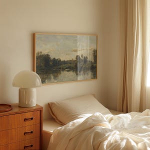 May include: A white bedside lamp with a white lampshade sits on a wooden dresser next to a bed with white linens. A framed landscape painting hangs on the wall behind the lamp.