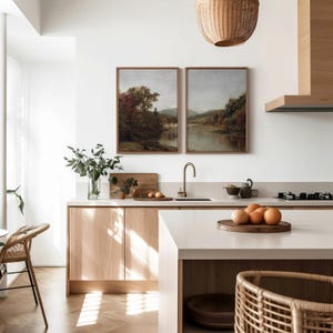 May include: A modern kitchen with white walls and light wood cabinets. Two framed landscape paintings hang above the kitchen sink. A wooden cutting board sits on the counter next to a bowl of fruit.