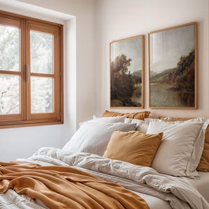 May include: A bedroom with a white bed with a brown blanket and a few pillows. There are two framed landscape paintings on the wall above the bed.