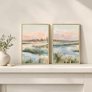 Puede incluir: Dos cuadros rectangulares con marcos dorados finos que presentan un paisaje de acuarela de una marisma al atardecer. La obra muestra tonos suaves de rosa, azul y verde, representando canales de agua que serpentean entre campos de hierba.