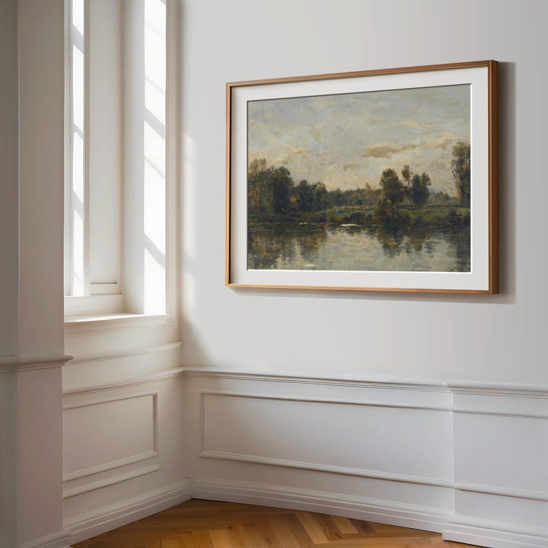 May include: A framed landscape painting depicting a river scene with trees and a cloudy sky. The painting is in a light wood frame and is hanging on a white wall.