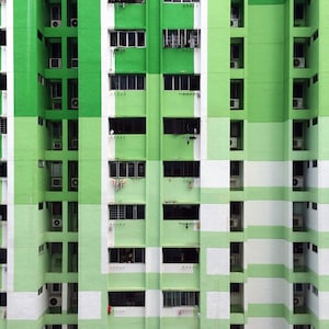 May include: A high-angle shot of a multi-story apartment building with a repeating pattern of green and white rectangular sections. Windows and air conditioning units are visible. Some windows have laundry hanging outside.