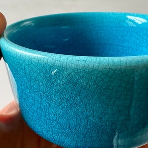 Puede incluir: Primer plano de un pequeño cuenco de cerámica turquesa con un esmalte agrietado texturizado. El interior y el exterior del cuenco son de un tono azul vibrante, con finas líneas visibles en toda la superficie. El cuenco está sostenido en una mano.
