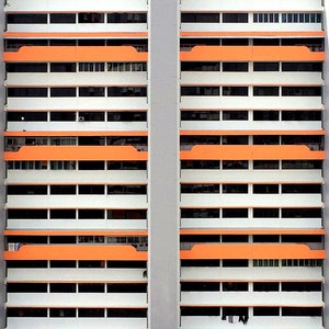 Op de afbeelding: Symmetrische weergave van een hoogbouwflatgebouw. Het gebouw heeft een herhalend patroon van witte muren, oranje balkons en donkere ramen. Verticale grijze steunbalken lopen langs de zijkanten van het gebouw.